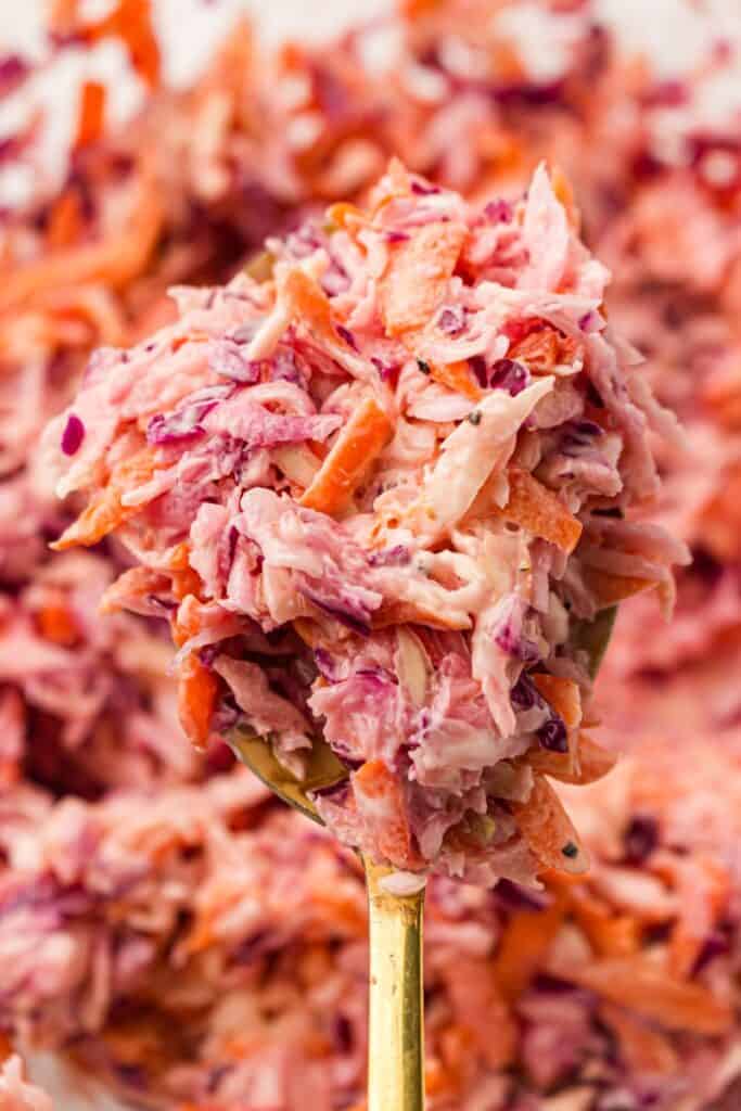 A close-up of a spoonful of colorful coleslaw with shredded red cabbage, carrots, and creamy dressing, with more coleslaw blurred in the background.