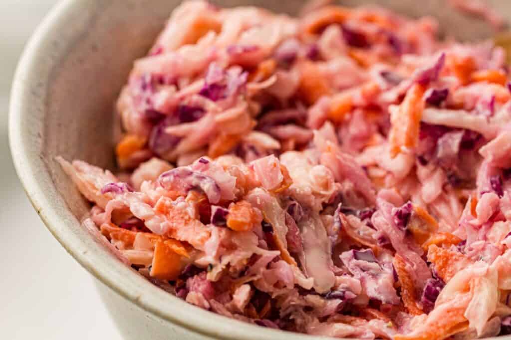 A close-up of creamy coleslaw in a bowl, showing shredded carrots, purple cabbage, and green cabbage mixed together.
