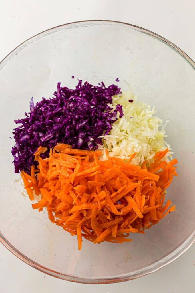 A glass bowl containing shredded purple cabbage, shredded green cabbage, and grated carrots, arranged in separate sections.
