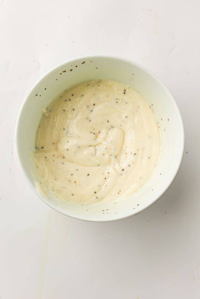 A white bowl filled with a creamy, speckled mixture sits on a light surface. The mixture appears to be seasoned with black pepper or herbs and is smooth and slightly thick in texture.