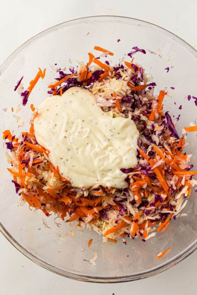 A glass bowl filled with shredded red cabbage, carrots, and white cabbage, topped with a dollop of creamy dressing, ready to be mixed for coleslaw.