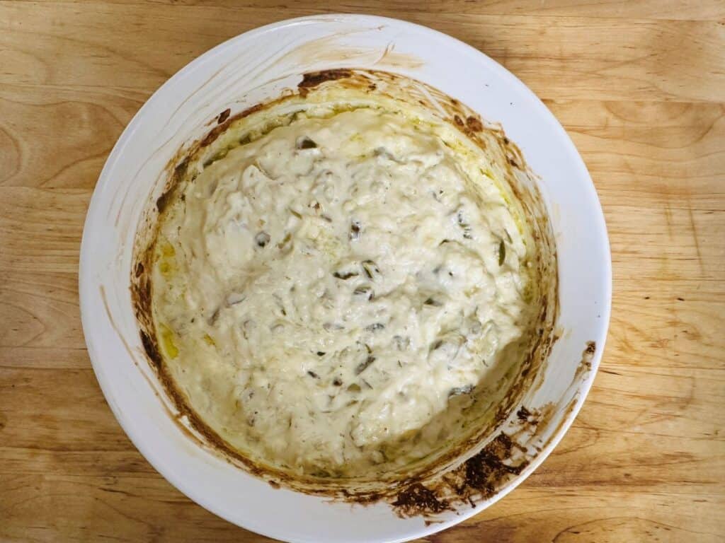 A round white dish with creamy, baked spinach artichoke dip. The edges of the dish have browned, baked-on cheese and sauce, and the dip has a slightly golden, bubbly surface. The dish sits on a wooden surface.