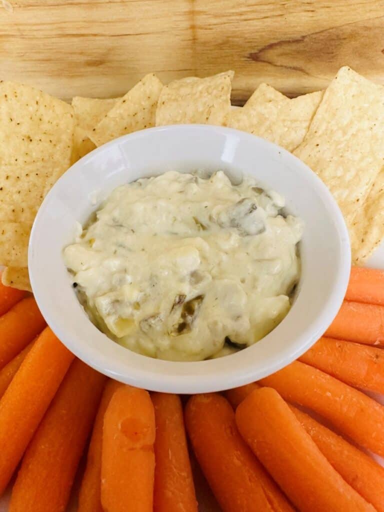 A white bowl of creamy dip is surrounded by tortilla chips and baby carrots, arranged in a semi-circle on a wooden surface.