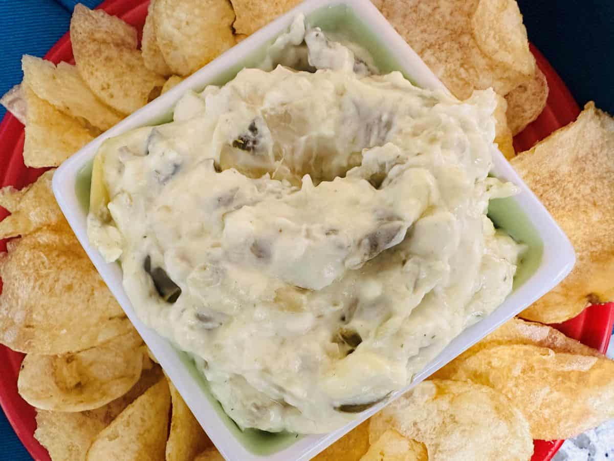 A bowl of creamy onion dip sits in the center of a red plate, surrounded by crispy potato chips.