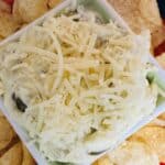 A square bowl filled with creamy dip topped with shredded cheese is surrounded by potato chips on a red plate.