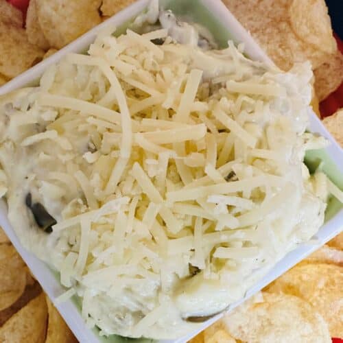 A square bowl filled with creamy dip topped with shredded cheese is surrounded by potato chips on a red plate.