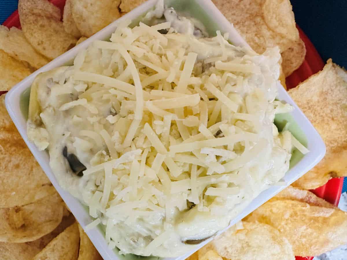 A square bowl filled with creamy dip topped with shredded cheese is surrounded by potato chips on a red plate.
