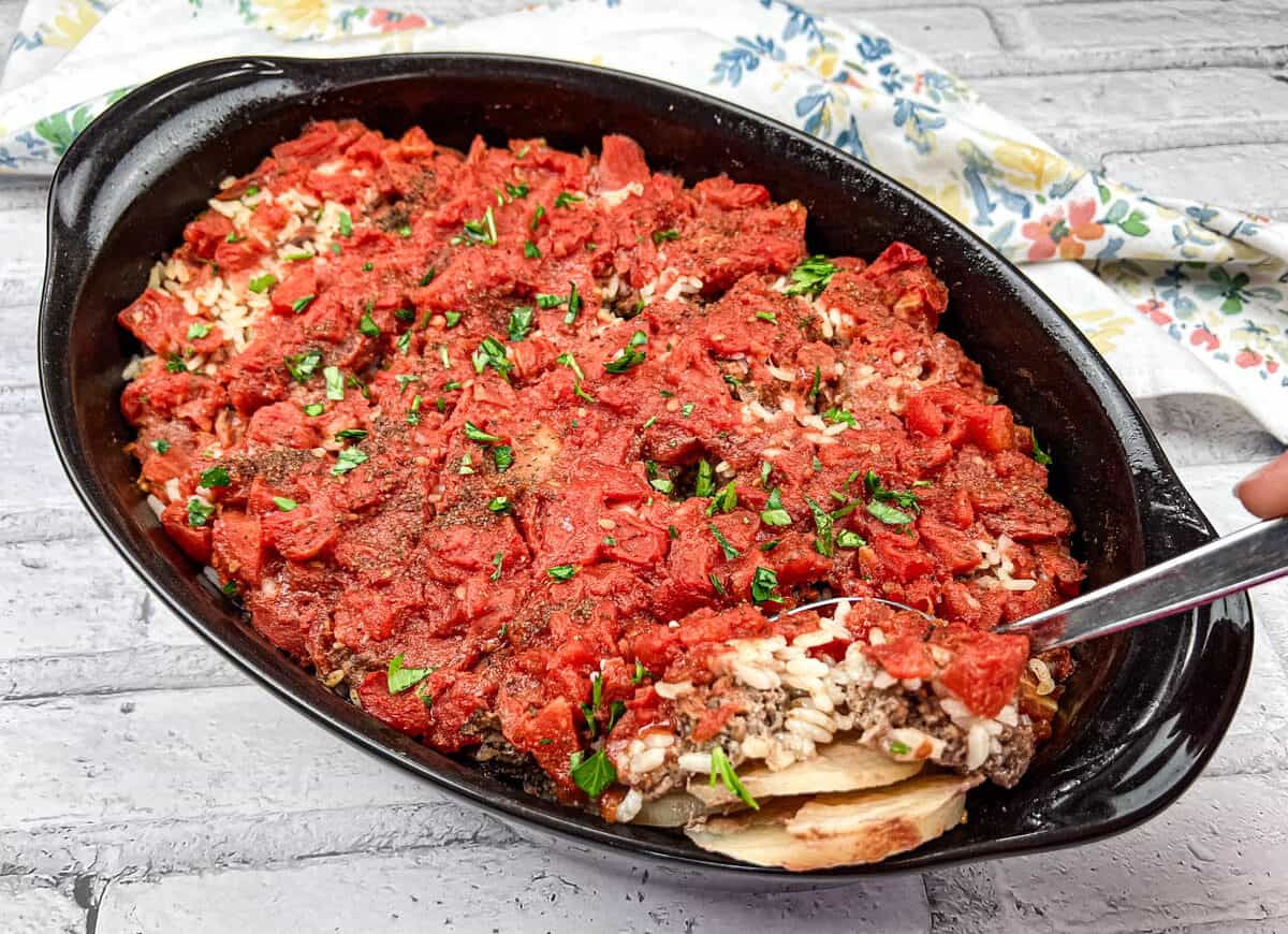 An oval black baking dish filled with a baked rice and meat casserole topped with tomato sauce and chopped herbs. A serving spoon is scooping out a portion. A floral cloth is nearby on a white brick surface.