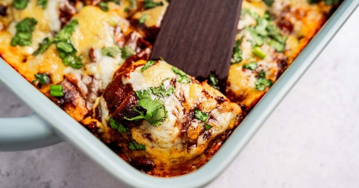 A close-up of cheesy enchiladas topped with chopped cilantro in a baking dish, with a spatula lifting one serving out.