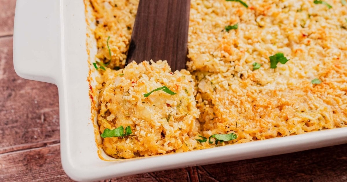A close-up of a baked casserole in a white dish, topped with golden breadcrumbs and garnished with chopped parsley. A spatula is lifting a serving from the corner, revealing the creamy filling.