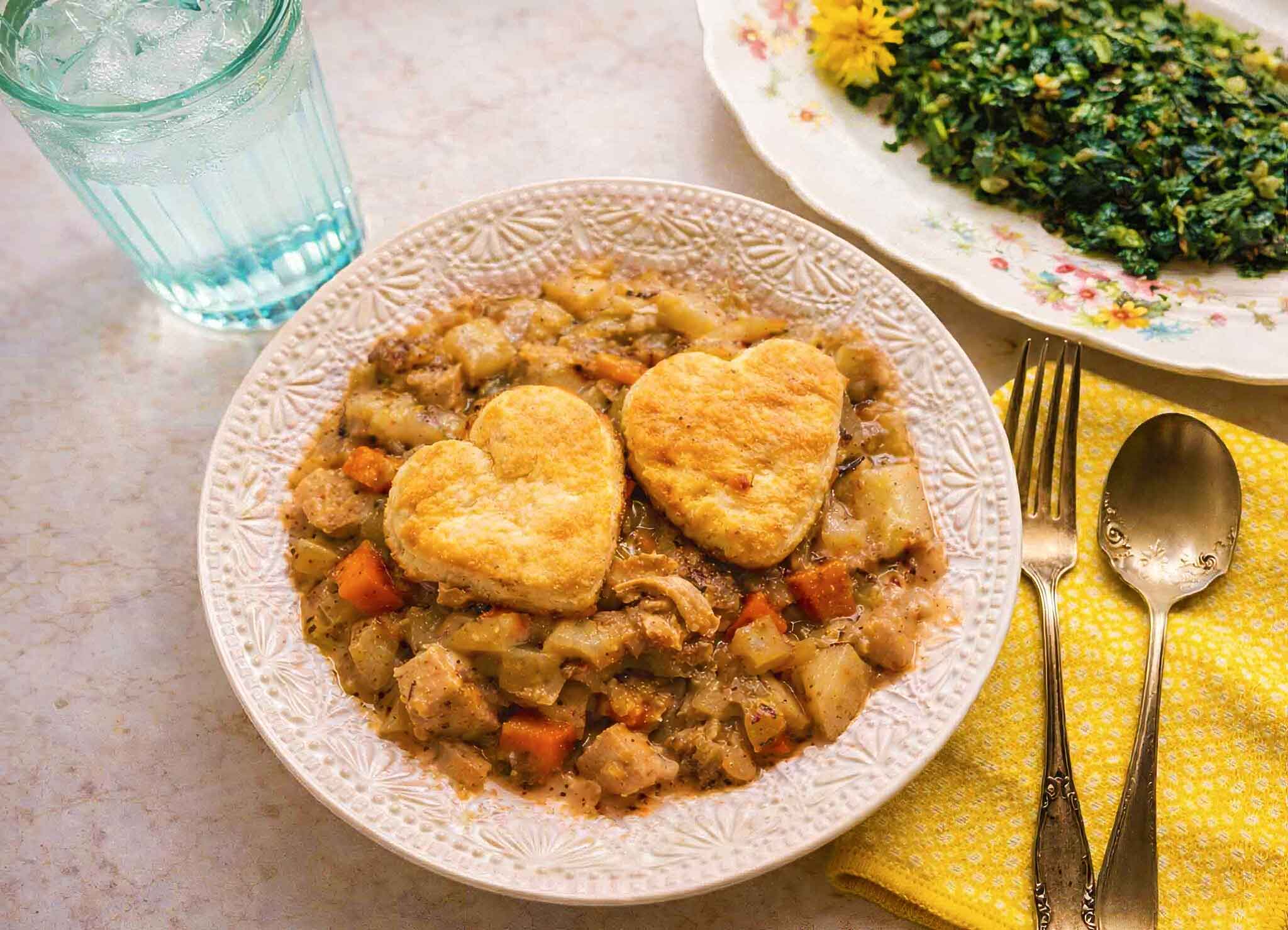 A bowl of stew with diced vegetables and meat, topped with two heart-shaped biscuits. Next to it is a glass of ice water and a plate of greens. A yellow napkin holds a spoon and fork.