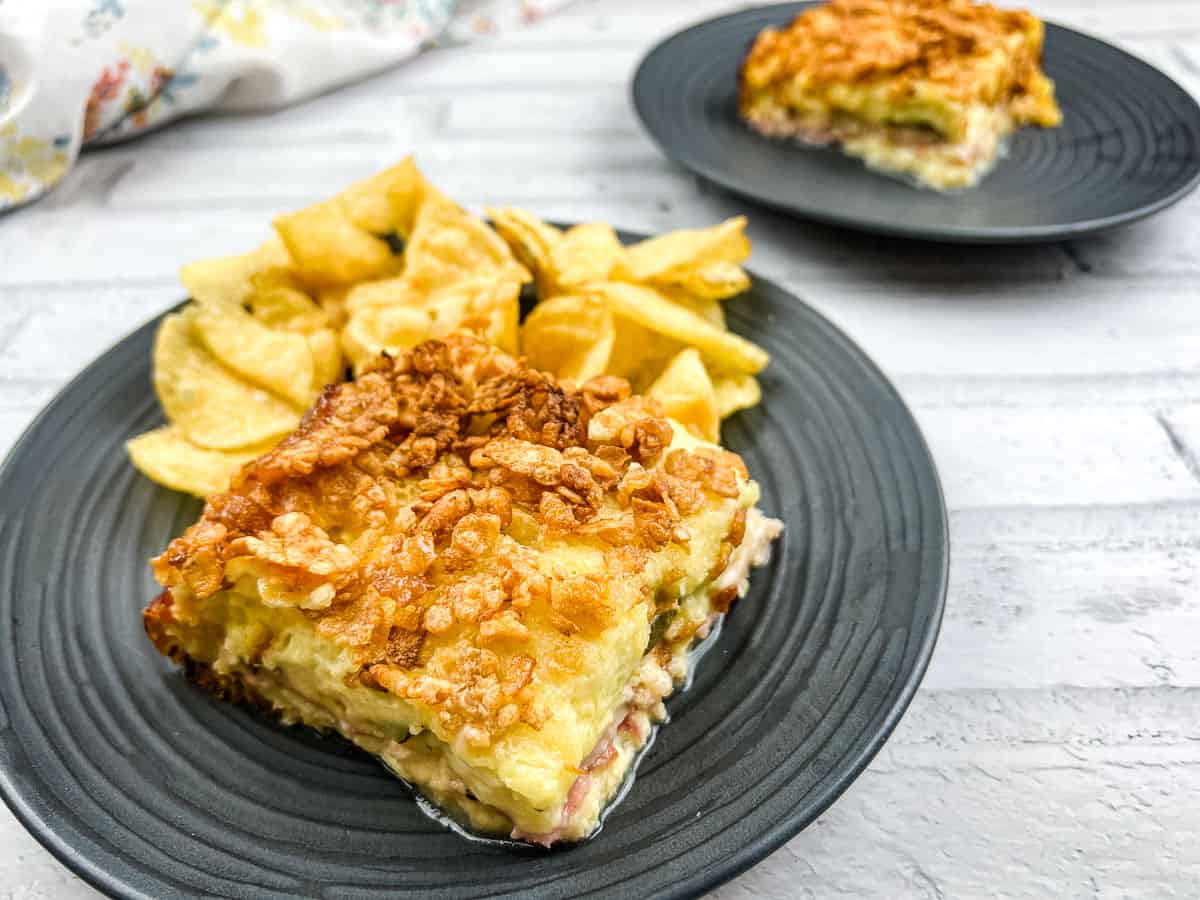 A black plate with a slice of savory casserole topped with a crispy golden layer, served with a side of potato chips. Another similar plate sits in the background.