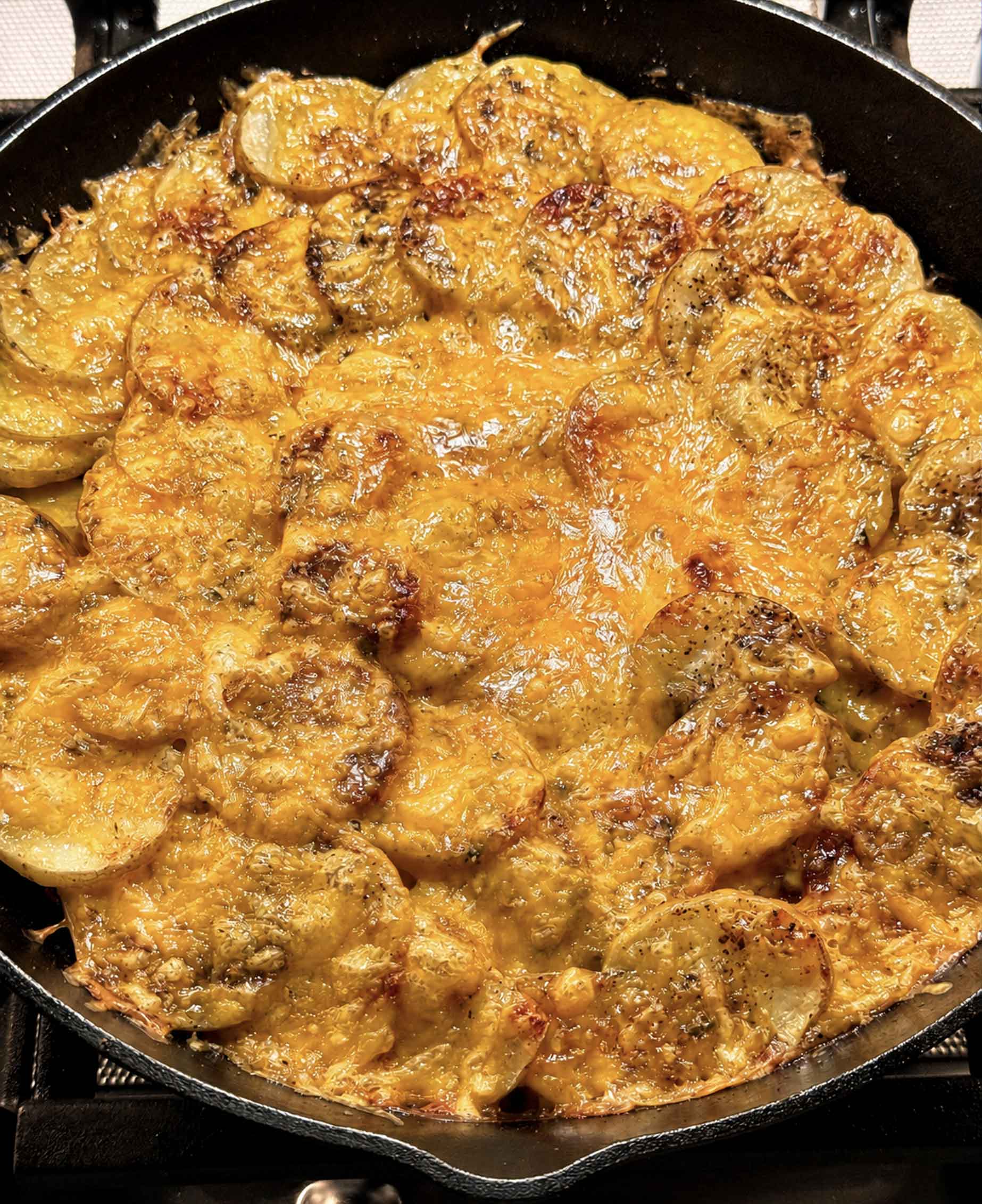 A close-up of a cast iron skillet filled with golden-brown, cheesy scalloped potatoes, baked with a crispy, bubbly top layer.