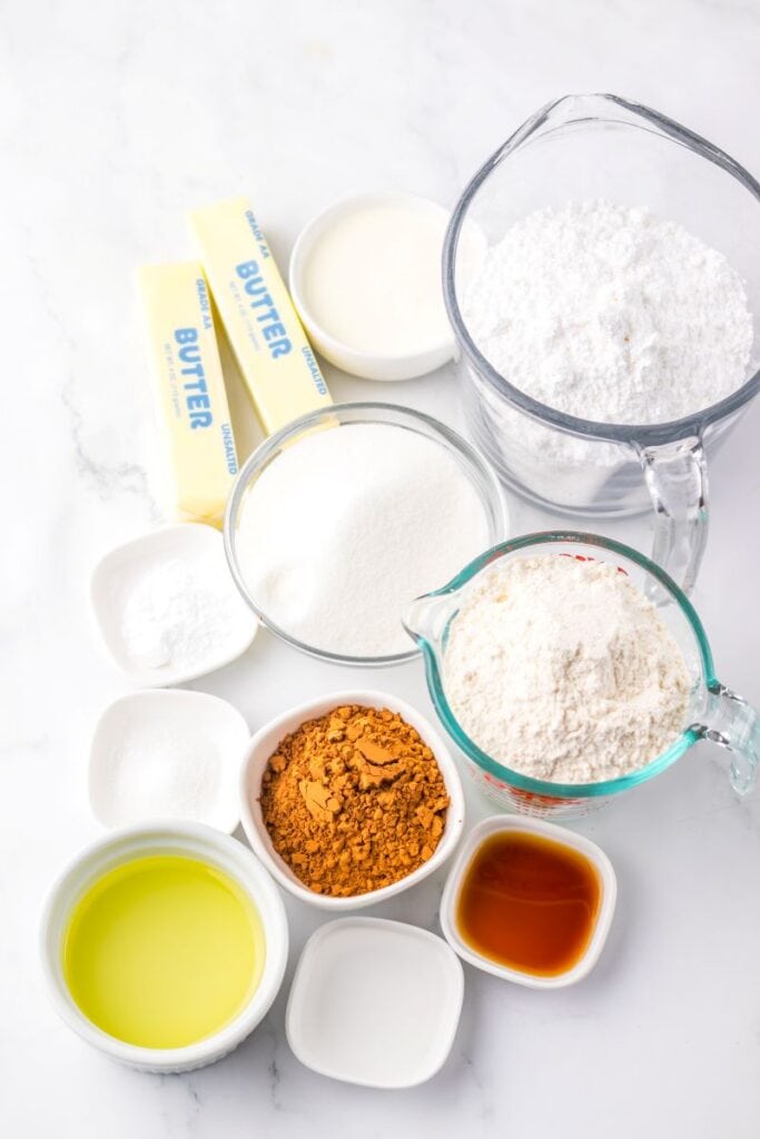 Various baking ingredients are arranged on a white surface, including butter sticks, white sugar, flour, powdered sugar, cocoa powder, vanilla extract, oil, and small bowls with other baking essentials.