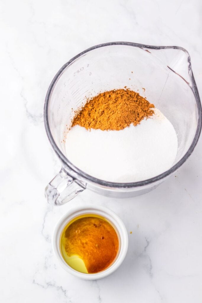 A glass measuring cup contains white sugar and cocoa powder. Beside it, a small white bowl holds golden liquid, likely oil or vinegar, all on a white marble surface.