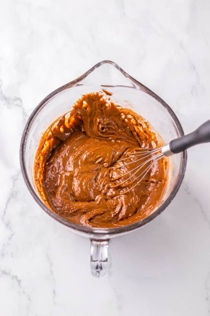 Glass mixing bowl with thick, brown batter and a whisk on a white marble surface. Some batter is visible on the sides of the bowl.