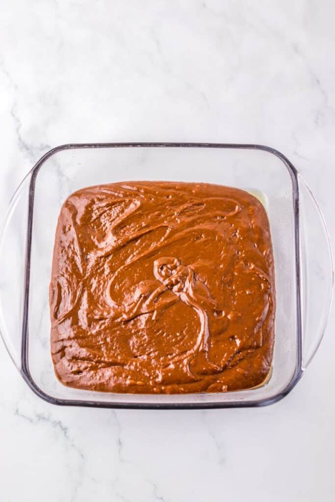 A glass baking dish filled with smooth, swirled chocolate brownie batter sits on a white marble surface.