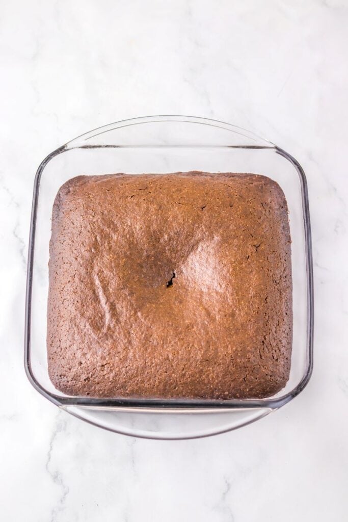 A square chocolate cake with a crack in the center sits in a clear glass baking dish on a white marble surface.