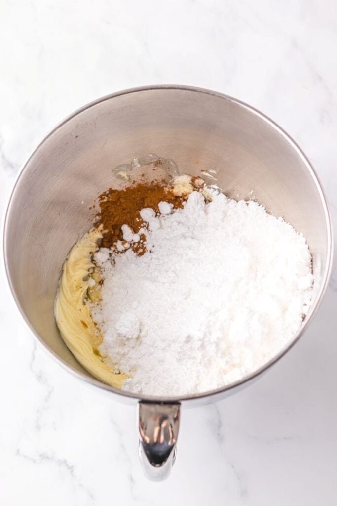 A metal mixing bowl contains creamed butter, powdered sugar, and brown spices on a white marble surface, ready to be mixed together.