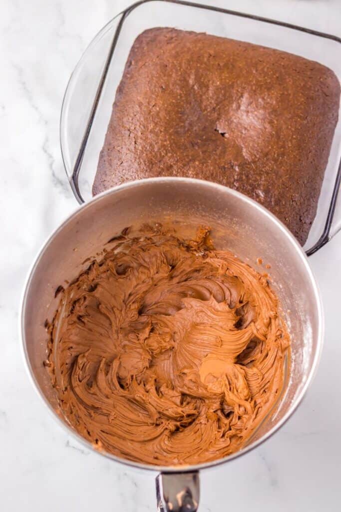 A bowl of chocolate frosting sits next to a baked chocolate cake in a glass dish on a marble surface, ready for icing.