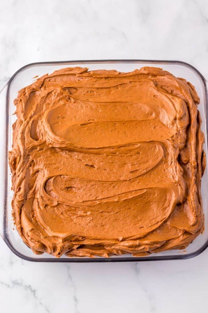 A glass baking dish filled with a square chocolate cake topped with swirled chocolate frosting, set on a white marble surface.