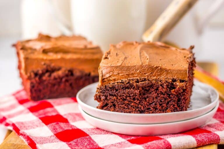 Two squares of chocolate cake with chocolate frosting on white plates, sitting on a red and white checkered cloth. A glass of milk is blurred in the background.