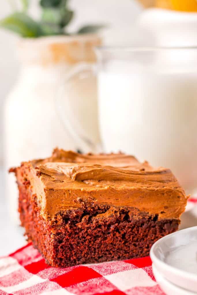 A close-up of a chocolate frosted brownie sits on a red and white checkered cloth, with a plate, a glass of milk, and a jar in the blurred background.