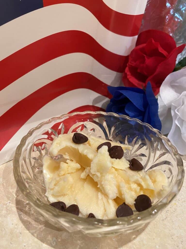 A glass bowl of vanilla ice cream with chocolate chips sits in front of an American flag decoration and red, white, and blue artificial flowers.