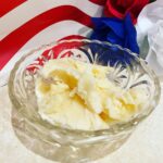 A glass bowl with scoops of vanilla ice cream sits on a beige surface. In the background, there are red, white, and blue decorative flowers and a partial view of a red stripe.