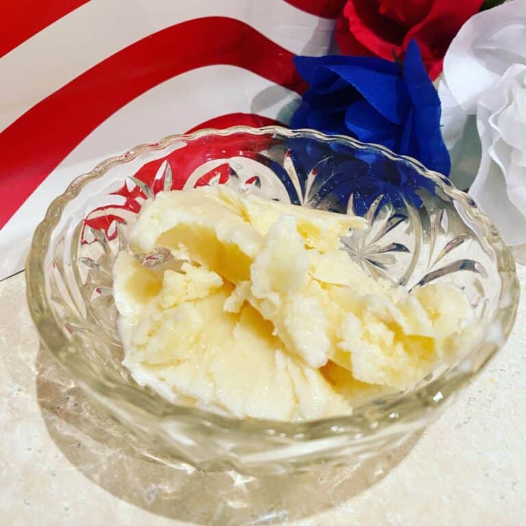 A glass bowl with scoops of vanilla ice cream sits on a beige surface. In the background, there are red, white, and blue decorative flowers and a partial view of a red stripe.