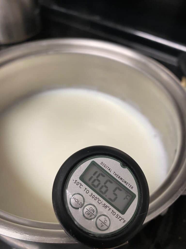 A digital thermometer reads 165.5°F while inserted into a pot of milk, indicating the milk is being heated.