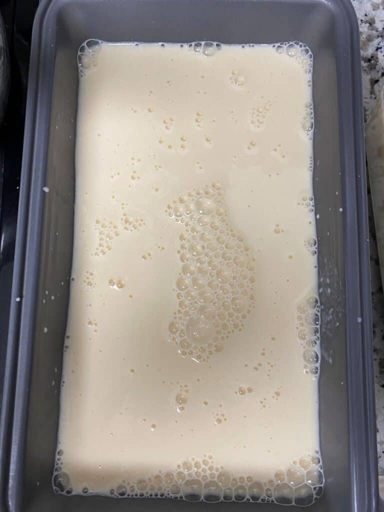A rectangular metal pan filled with a pale, creamy liquid topped with small bubbles, sitting on a granite countertop.