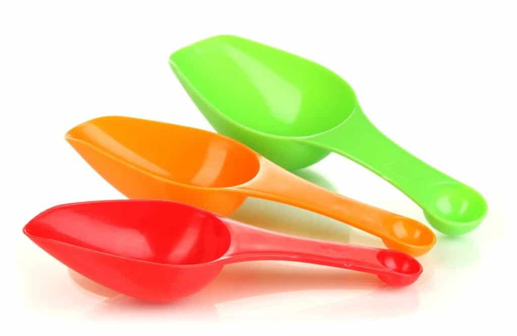 Three plastic scoops in red, orange, and green are stacked on top of each other against a white background. The handles are aligned, with the green scoop on top, followed by orange, and then red.