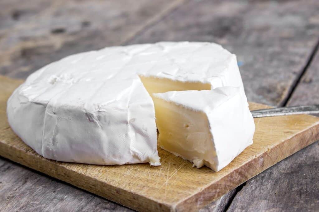 A wheel of soft white cheese, likely Brie or Camembert, with a wedge cut out and placed beside it on a wooden cutting board. The cheese has a creamy interior and a white rind.