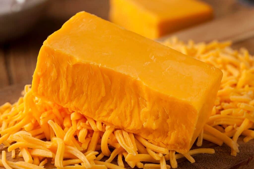 A block of cheddar cheese rests on a pile of shredded cheddar cheese on a wooden surface, with another block of cheese blurred in the background.