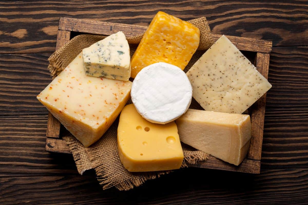 A wooden tray lined with burlap holds an assortment of cheeses, including wedges and rounds of various types, colors, and textures, all displayed on a dark wood table.