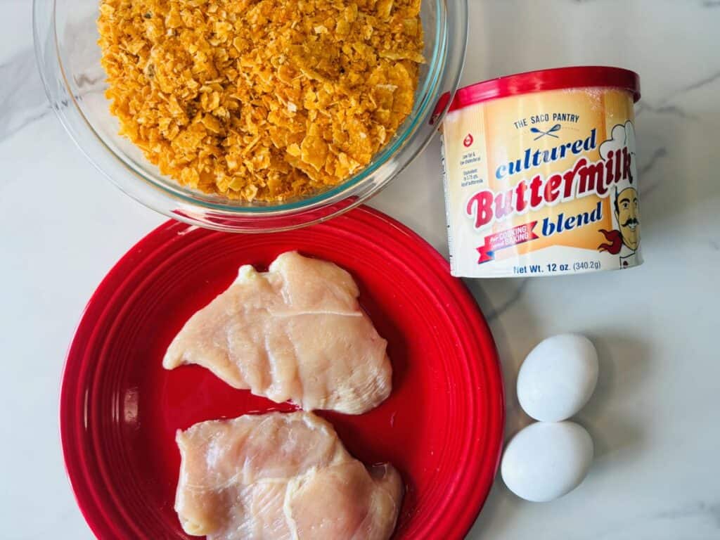 A bowl of crushed cornflakes, a can of cultured buttermilk blend, two eggs, and two raw chicken breasts on a red plate are arranged on a white surface.