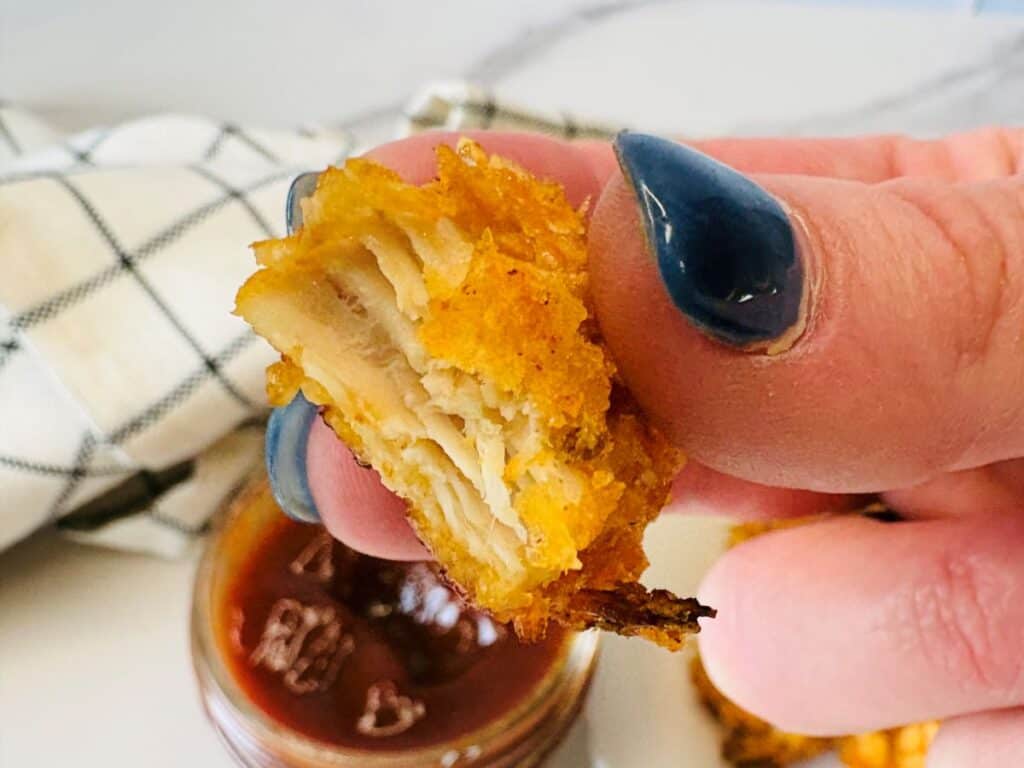A hand with blue-painted nails holds a crispy, golden-brown, layered bite of fried food above a dish of dipping sauce, with a checkered napkin in the background.