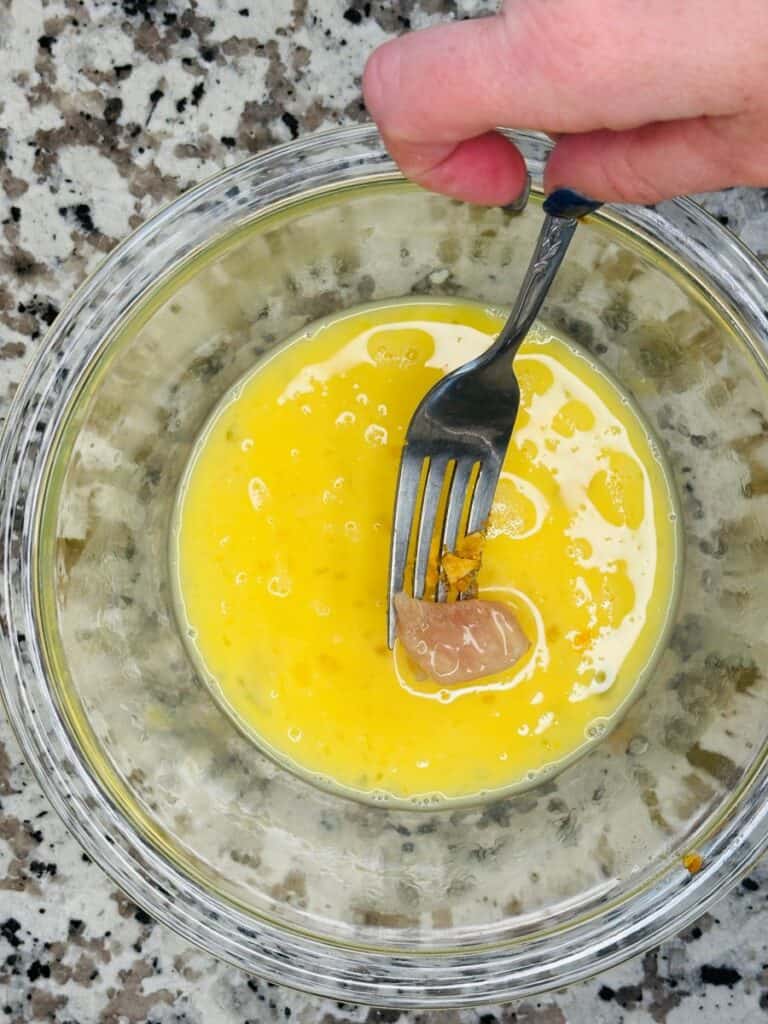 A hand holding a fork mixes cracked eggs with a piece of raw sausage in a glass bowl on a speckled countertop.