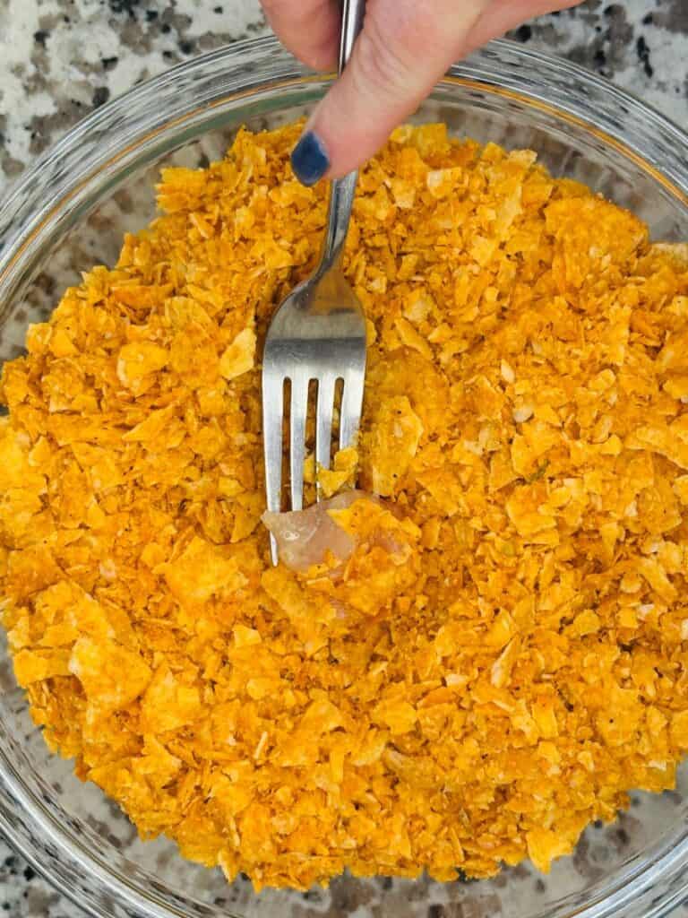 A hand holding a fork presses a small piece of raw chicken into a bowl filled with crushed potato chips on a granite countertop.