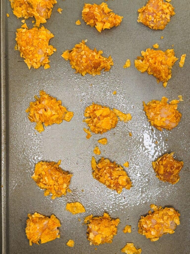 Small mounds of orange, crumb-covered snacks are arranged on a greased metal baking sheet, spaced evenly in rows and columns, ready to be baked.