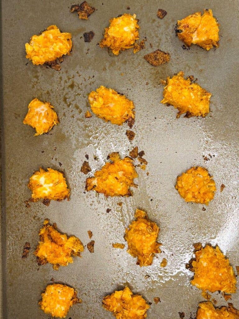 Small, crispy, golden-brown baked nuggets are scattered across a greased baking sheet. The nuggets appear textured and irregular in shape, with some browned edges and oily spots on the pan.