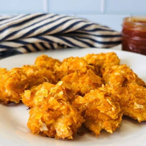 A white plate holds several crispy, golden-brown chicken nuggets with a jar of dipping sauce and a blue-and-white striped cloth in the background.