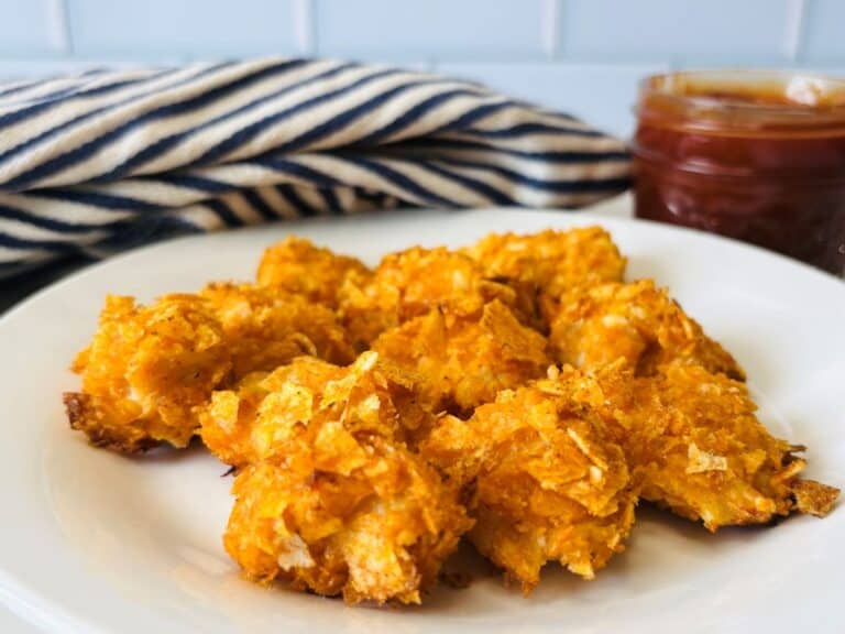 A white plate holds several crispy, golden-brown chicken nuggets with a jar of dipping sauce and a blue-and-white striped cloth in the background.