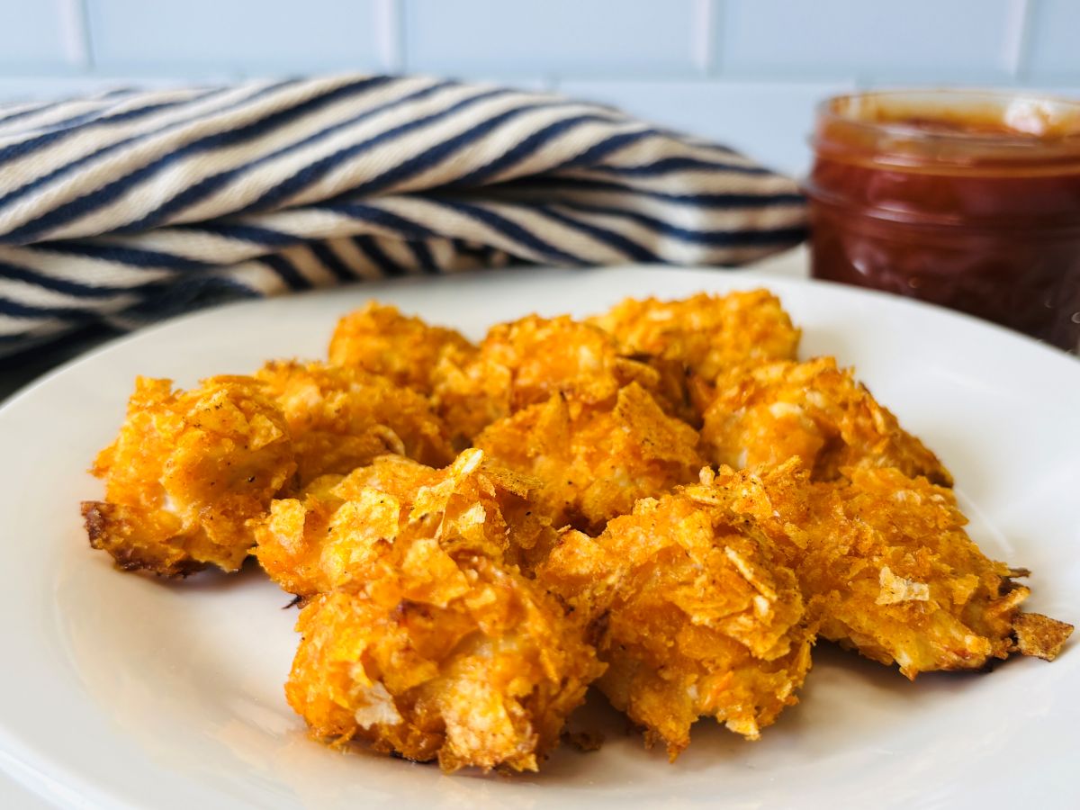 A white plate holds several crispy, golden-brown chicken nuggets with a jar of dipping sauce and a blue-and-white striped cloth in the background.
