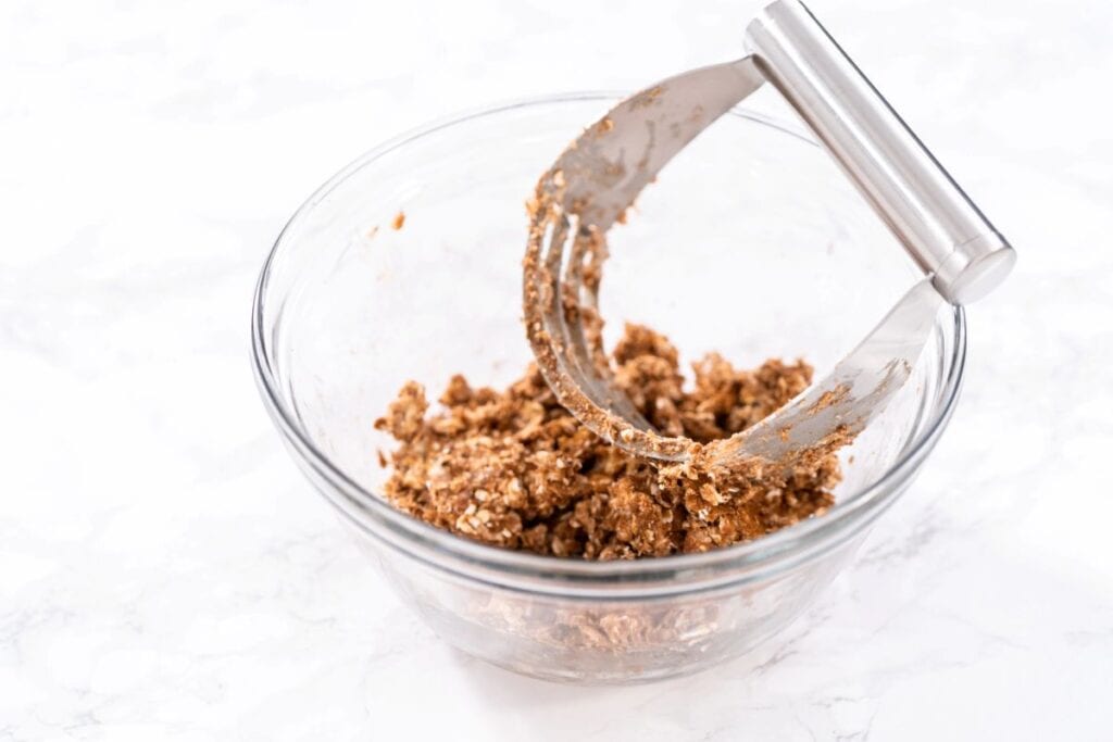 A glass bowl containing a chunky, brown dough mixture is shown with a stainless steel pastry cutter resting inside.