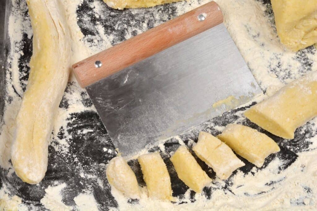 A dough scraper rests on a floured surface beside a partially sliced roll of dough, with several cut pieces scattered around, ready for further preparation.