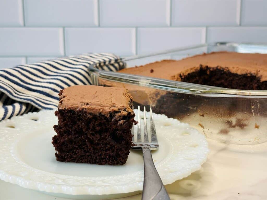 A slice of chocolate cake with frosting on a white plate and fork, with the rest of the cake in a glass pan in the background, next to a striped cloth.