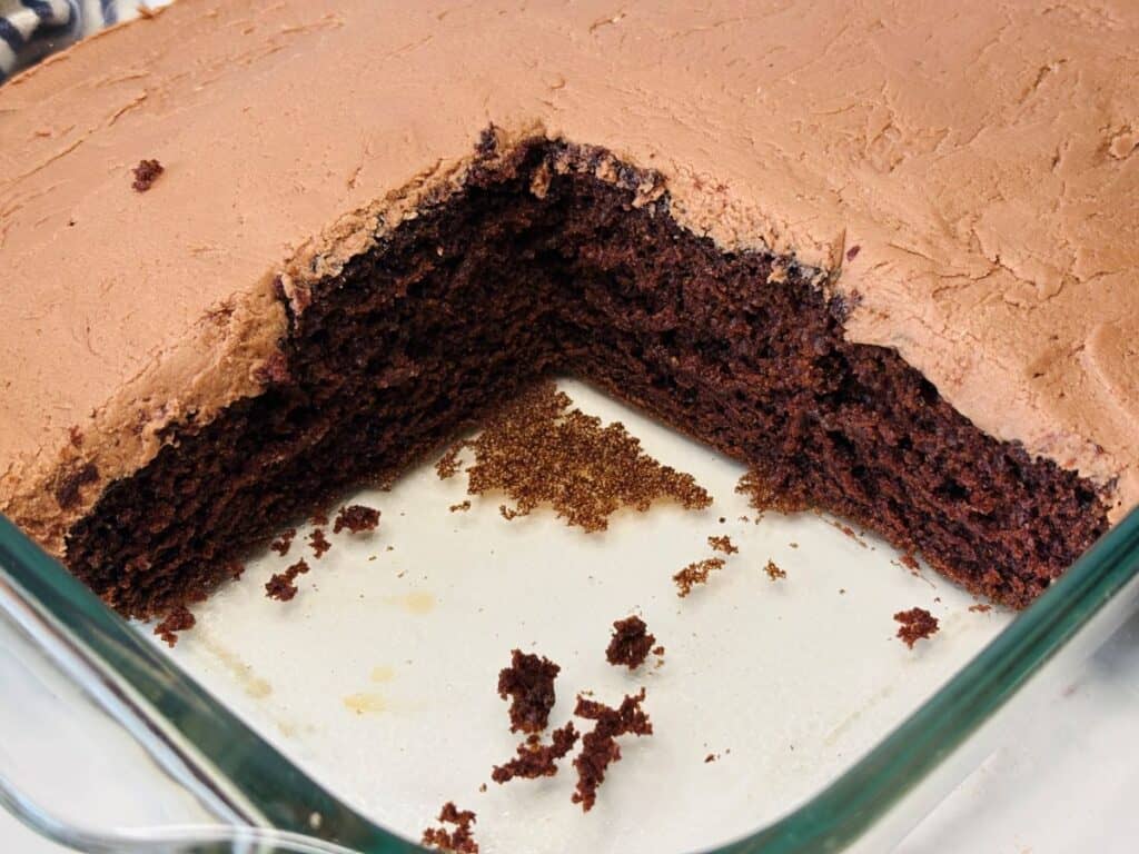 A rectangular chocolate cake in a glass dish with a large square piece missing, exposing the moist, dark interior and fluffy chocolate frosting on top. Crumbs are scattered in the empty corner.