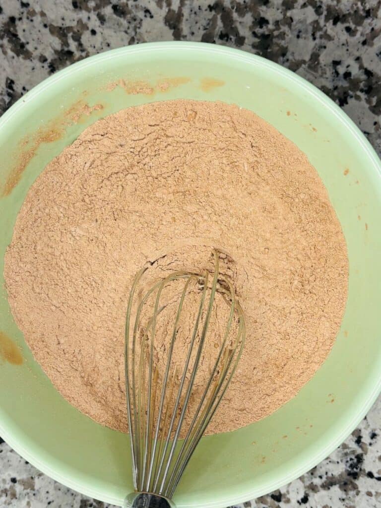 A green bowl filled with light brown cocoa powder mixture is being whisked with a metal whisk. The bowl sits on a granite countertop.
